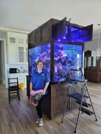 A person stands next to a large, illuminated aquarium in a room with wooden floors and white cabinets. A step ladder and a yellow bucket are nearby.